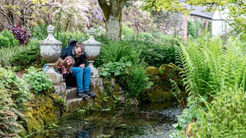 An adult and child sitting on stone steps next to a small stream, surrounded by lush foliage
